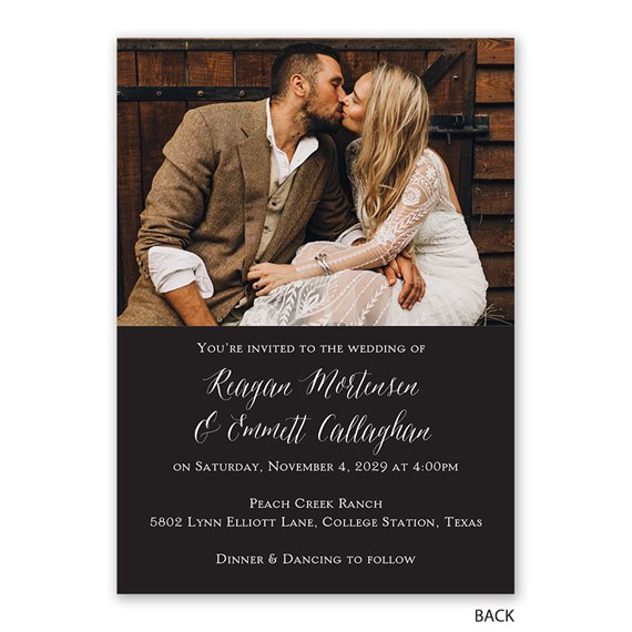 Photo wedding invitation with bride in lace dress and groom in brown tweed suit kissing in front of a wooden wall; dark lower panel with white script text.