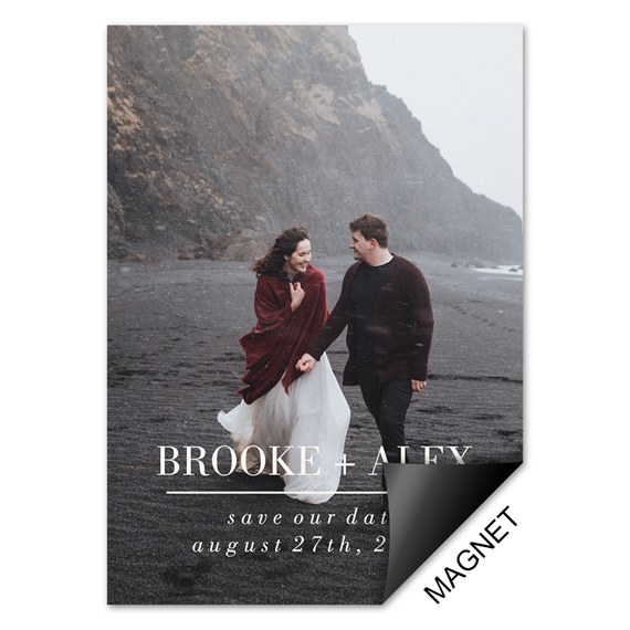 Save-the-date photo magnet with couple on a black sand beach, woman in white dress and burgundy shawl and man in dark sweater, white printed names and date