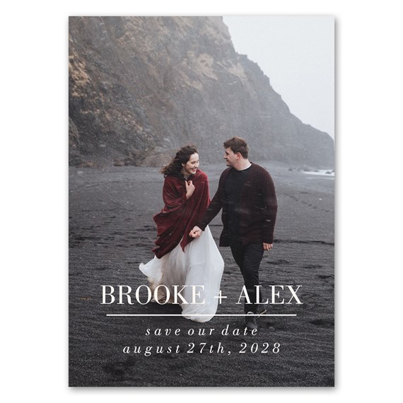Couple walking on black sand beach for save-the-date magnet; woman in white dress with burgundy shawl and man in dark sweater with white text overlay