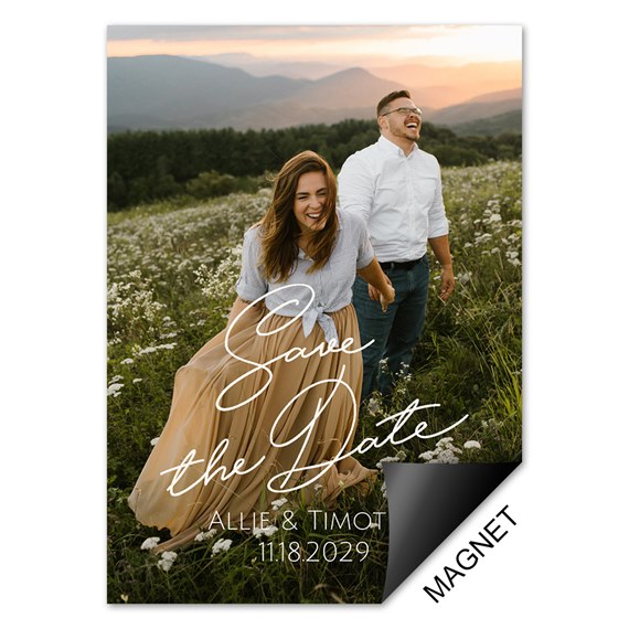 Save-the-date photo magnet of a couple in a wildflower field at sunset with white script text overlay.