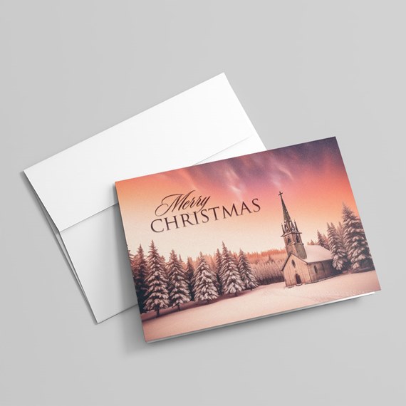 Folded Christmas card and white envelope showing a snowy forest and church under a pink-orange sky with 'Merry Christmas' text