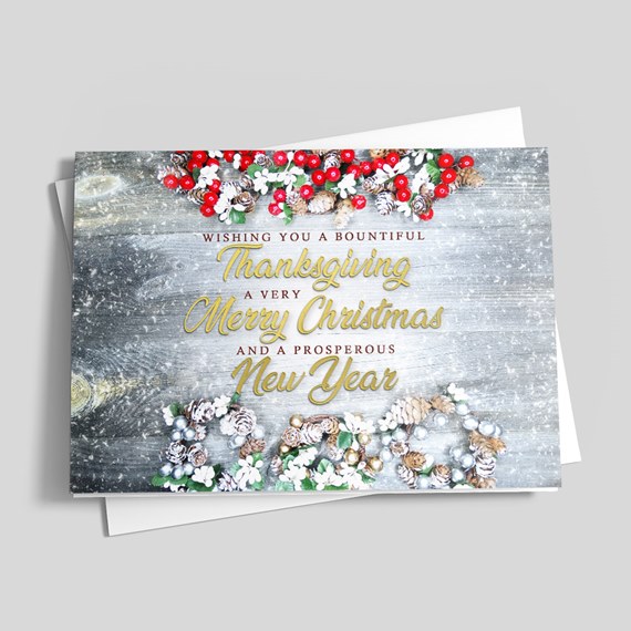 Holiday greeting card with gold script on gray wood background, decorated with red berries, pinecones, white flowers and silver ornaments, shown with a white envelope.