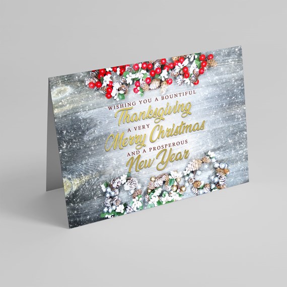 Folded holiday greeting card with pinecones, red berries and white flowers on a gray wood background with gold script holiday greeting.