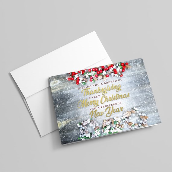 Folded holiday greeting card and white envelope on gray wood background, decorated with pinecones, red berries, greenery, snow accents and gold script text
