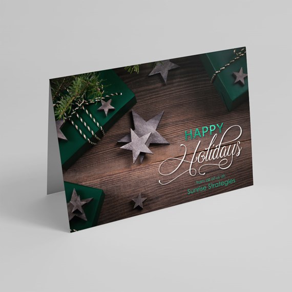 Folded holiday greeting card on wooden background with gray star ornaments, green wrapped gifts, twine, pine sprigs and scripted "Happy Holidays" text