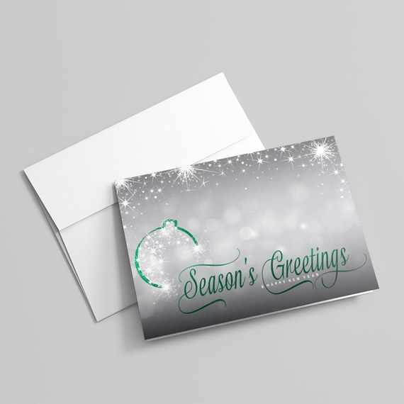 Folded holiday greeting card and white envelope with silver-gray sparkly background, green "Season's Greetings" script and ornament outline