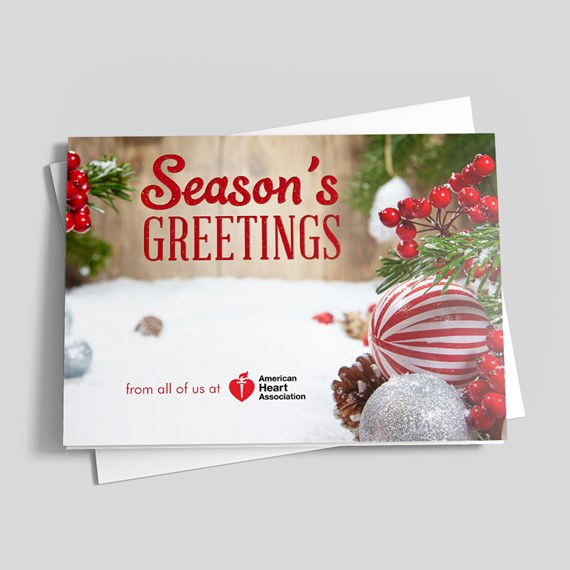 Holiday greeting card with Season's Greetings text, red striped ornament, silver bauble, pine branches and red berries on snowy wood background