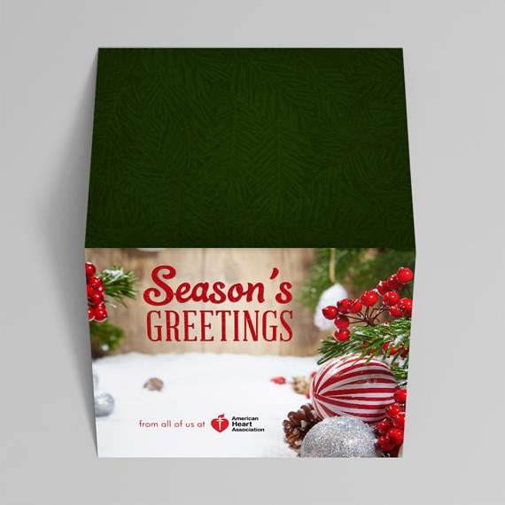 Holiday gift box with green lid and "Season's Greetings" front showing red berries, evergreen sprigs, pine cones, striped ornament and glitter ball on snow.