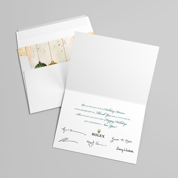 White folded holiday greeting card with green text and gold ornament liner displayed with white envelope