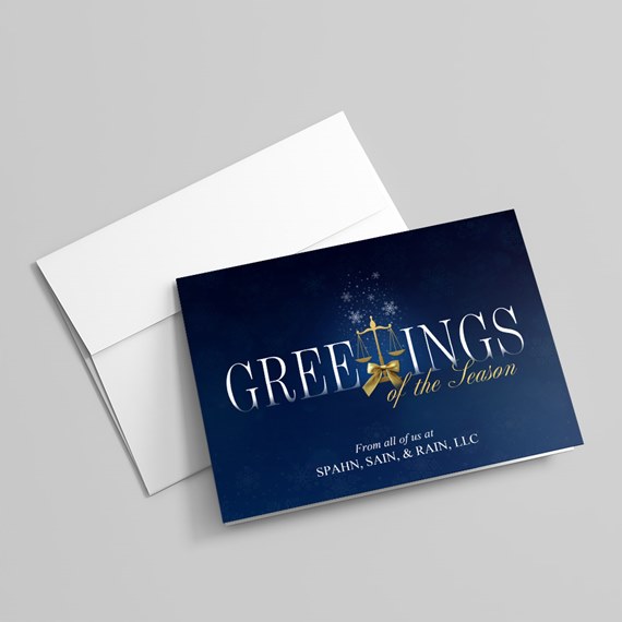 Folded navy holiday greeting card with white lettering and a gold scales-of-justice emblem, shown with a white envelope