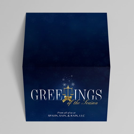 Folded navy holiday card with white 'Greetings of the Season' text, gold scales emblem and bow, and subtle snowflake pattern