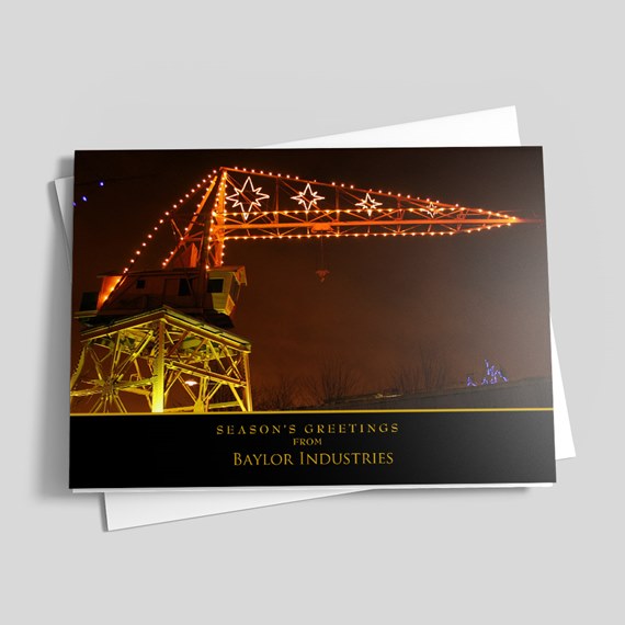 Holiday greeting card showing an illuminated industrial crane at night with string lights and star-shaped decorations, black lower band with gold stripe and seasonal greeting.