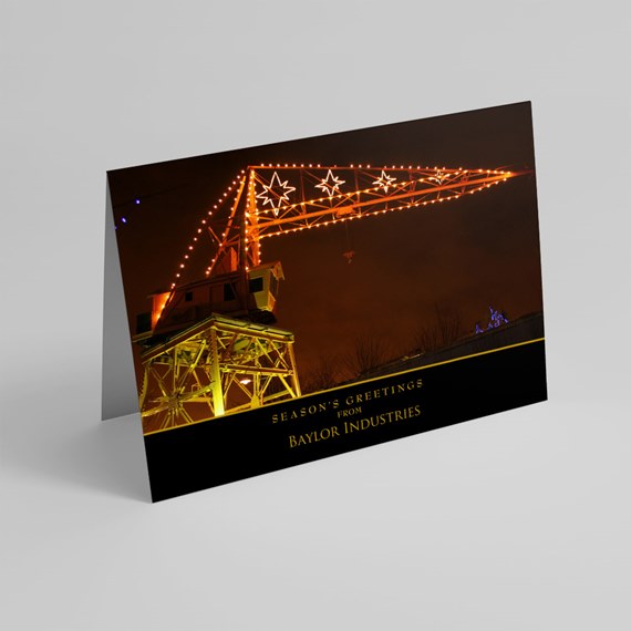 Folded holiday card with illuminated industrial crane at night, orange string lights, and black band with gold text.