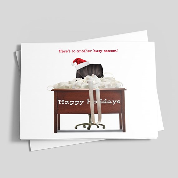 Holiday greeting card with wooden desk, piled papers, black office chair wearing a Santa hat and 'Happy Holidays' text