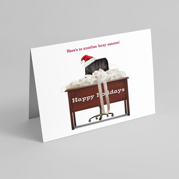 Folded holiday card showing an office desk covered in white paper streamers, a swivel chair wearing a Santa hat and 'Happy Holidays' printed on the desk.
