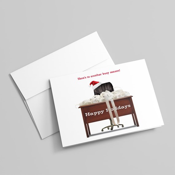 Folded holiday greeting card with office desk piled with papers, chair wearing a Santa hat and 'Happy Holidays' text, shown with a white envelope