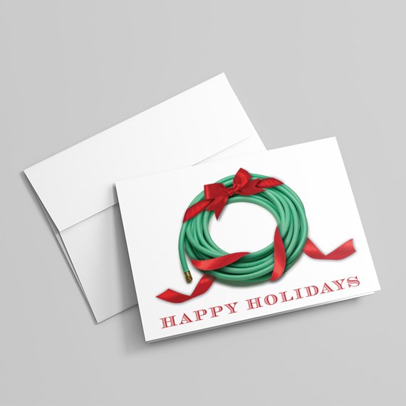 Folded holiday greeting card with a coiled green garden hose tied with a red ribbon forming a wreath, brass fitting visible and 'Happy Holidays' text; white envelope behind.