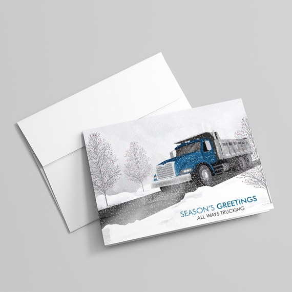 Folded holiday greeting card showing a blue dump truck on a snowy road with bare trees and falling snow, shown with a white envelope.