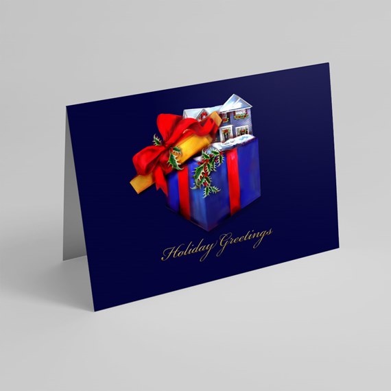 folded navy holiday greeting card with blue gift box, red bow, snowy house, holly accents and gold "Holiday Greetings" script