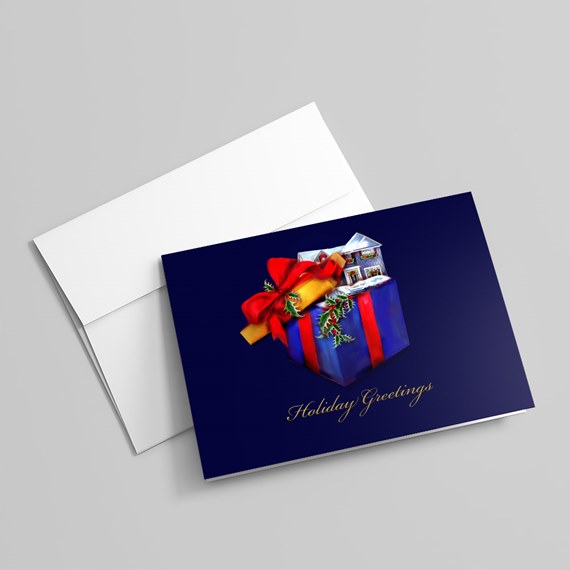 Folded navy holiday greeting card with illustrated gift box, red bow, snowy house and gold script, shown with a white envelope