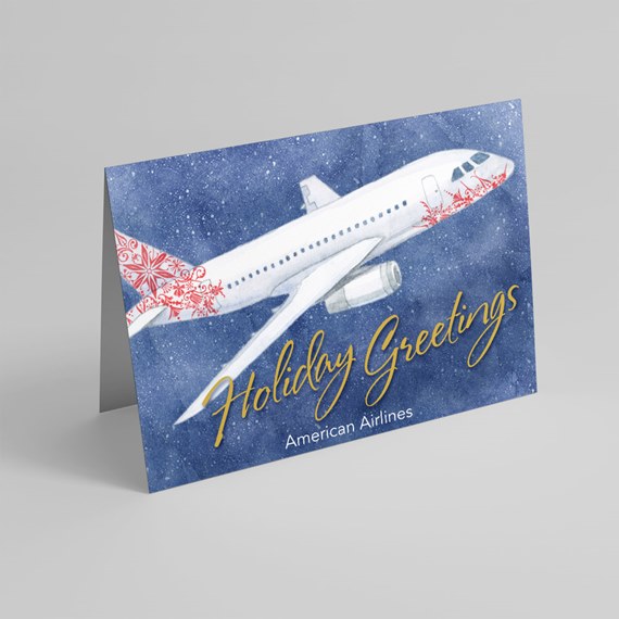 Folded holiday card with white airplane illustration, red ornament pattern and gold "Holiday Greetings" script on a blue snowy background.