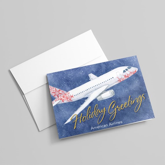 Holiday greeting card with white airplane illustration on a blue snowy background, gold script text and red decorative accents, shown with a white envelope.